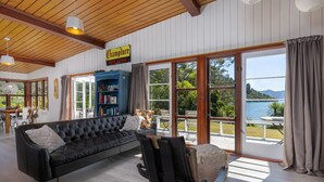 A cozy living room with a black leather sofa, a massage chair, and a bookshelf. Large windows offer a view of the outdoors. - Waterfront Marlborough Sounds Retreat 10min drive from Picton (Whatamango Bay)