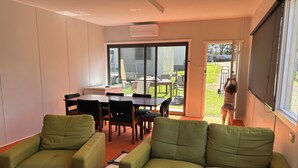 Una sala de estar con sofás verdes, una mesa de comedor y sillas. Hay un ventanal corredero que da a un área exterior con césped y mobiliario de patio.