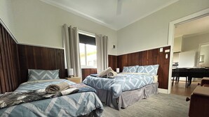 Habitación de hotel con dos camas, cabecero de madera, ventana con cortinas, una mesita con lámpara y una kitchenette al fondo.