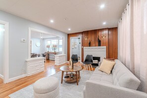 Ein modernes Wohnzimmer mit einer Couch, einem Hocker und einem runden Couchtisch. Ein Kamin mit Sims und eine Uhr an der Wand sind ebenfalls vorhanden.