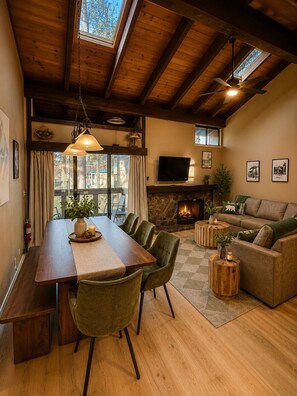 Una sala de estar acogedora con chimenea, un comedor con una mesa larga y sillas, un sofá y un televisor montado en la pared.