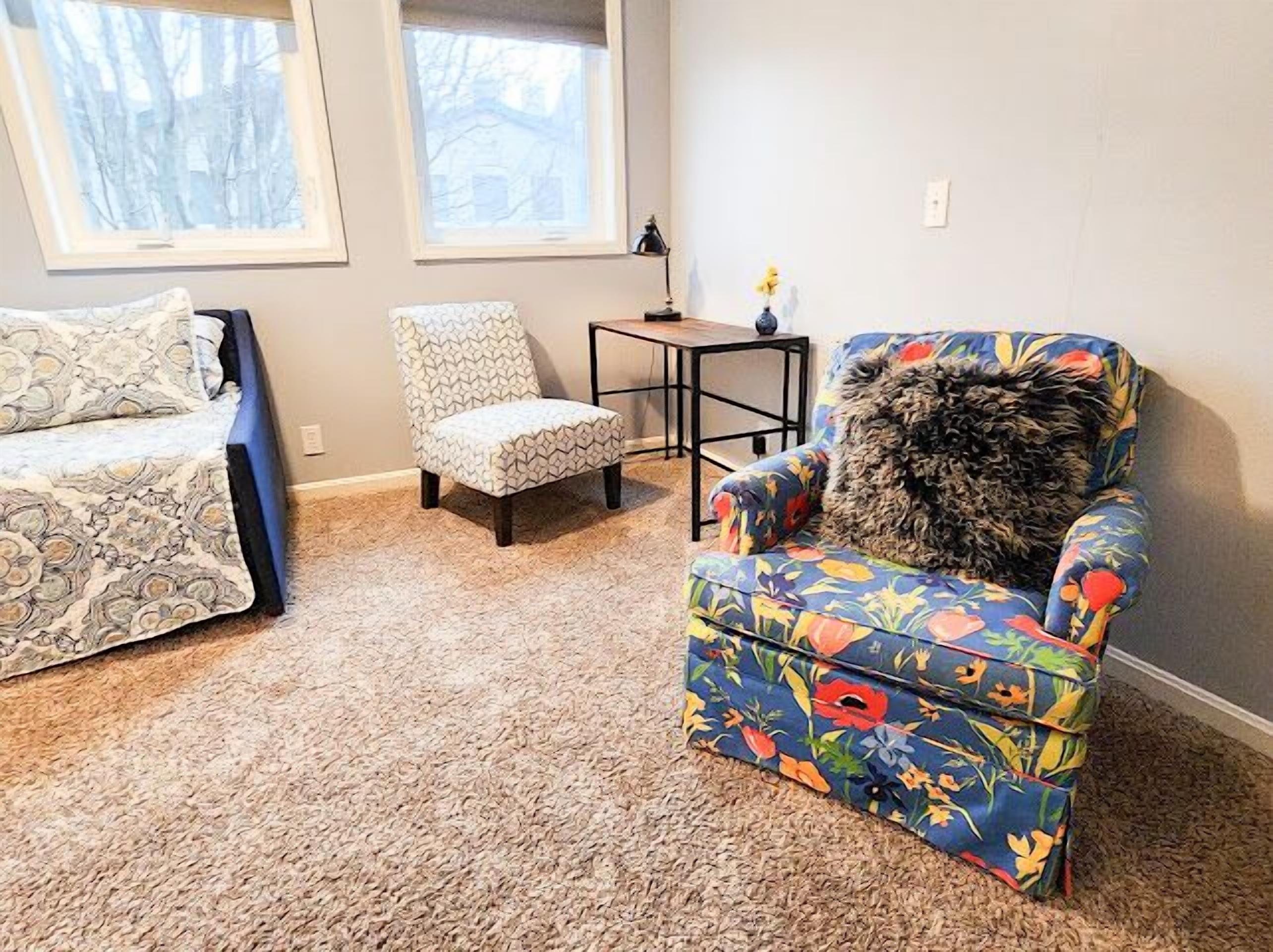 Una habitación acogedora con alfombra estampada, un sillón floral, un otoman a juego, una mesita y una ventana con vistas a árboles.