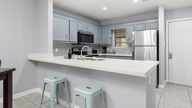 Una cocina moderna con armarios grises, encimera blanca, electrodomésticos de acero inoxidable y dos taburetes azul claro.