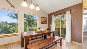 A dining area with a wooden table and bench, large windows with a view of trees, and a balcony with a railing. - Moose Mountain Lodge Above Park City (Summit County)