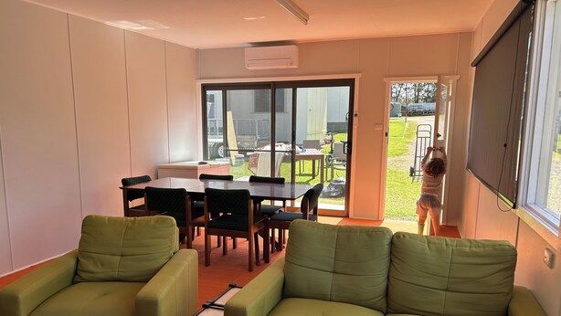 A room with a green sofa, a dining table with chairs, and a sliding glass door leading to an outdoor area with a lawn and patio furniture.