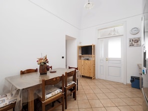 A dining area with a wooden table and chairs, a vase of flowers, a television on a wooden stand, and a kitchen area with a refrigerator and a clock on the wall. - Paravis Mio (Piano di Sorrento)