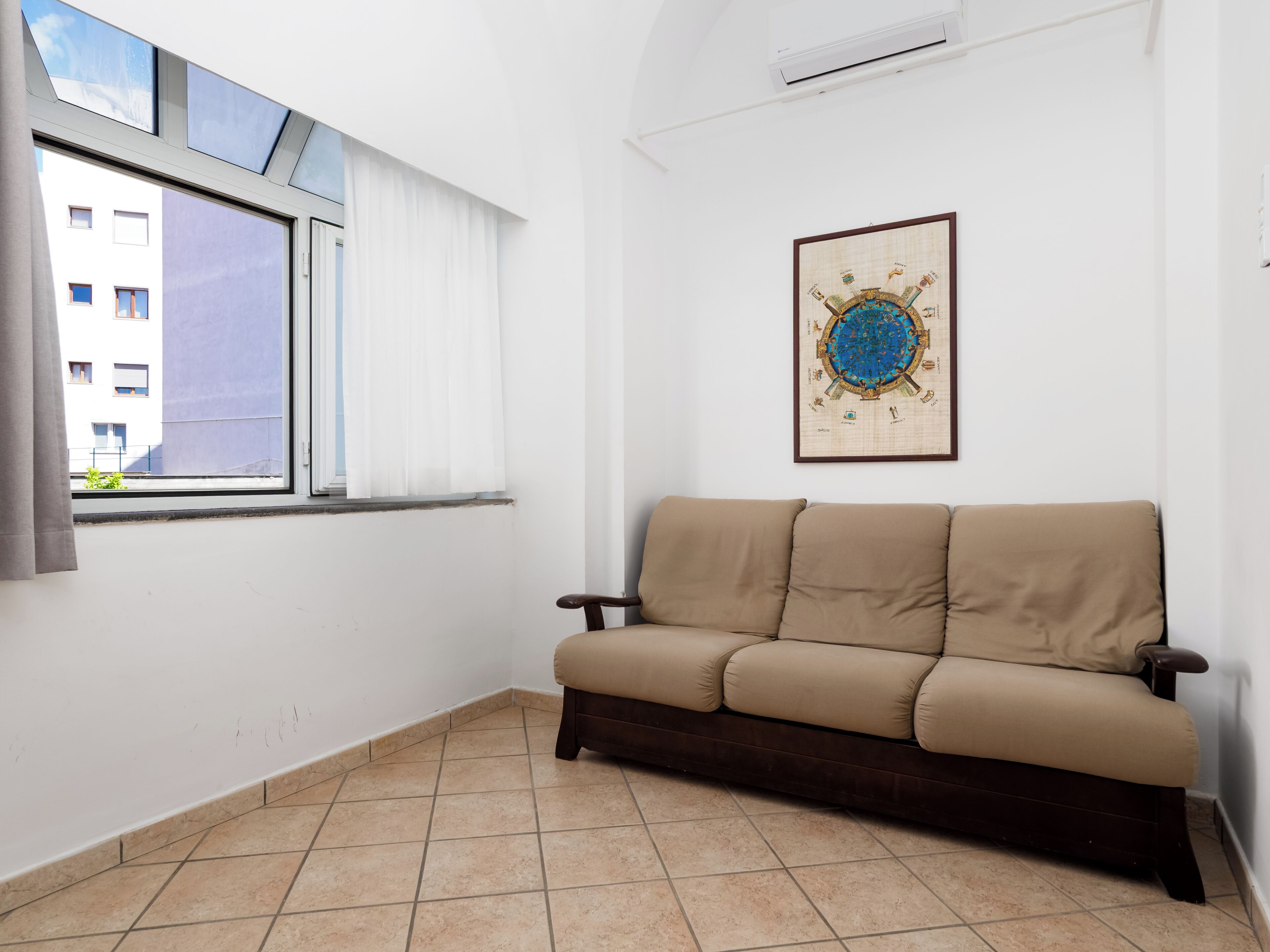 A room with a beige sofa, a window with white curtains, a wall-mounted air conditioner, and a framed map.