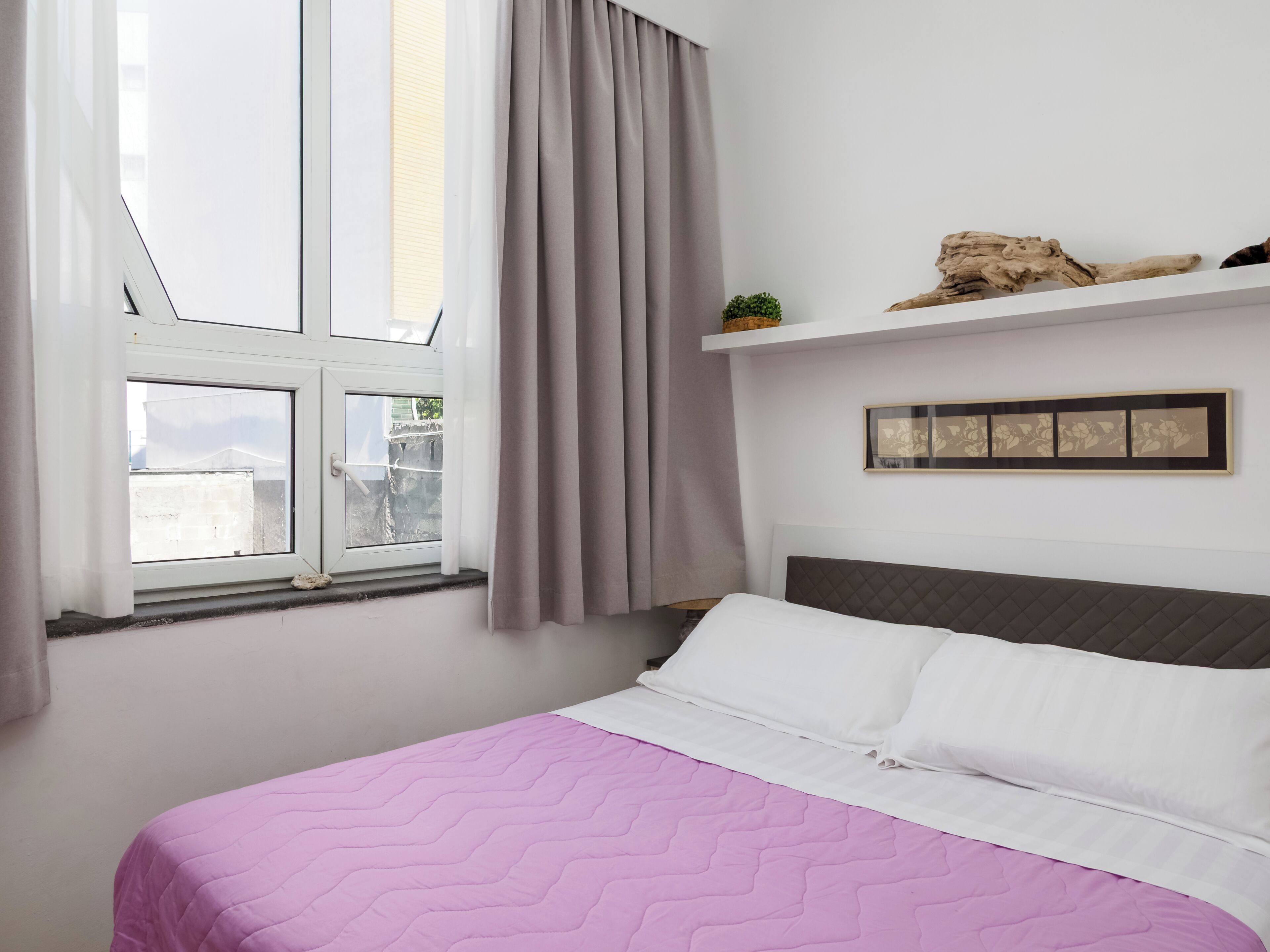 A bedroom with a bed, a window with curtains, a wall-mounted shelf with decorative items, and a framed picture.