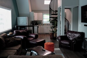 A modern living room with leather sofas, a coffee table, and a TV. A kitchen is visible in the background with white cabinets and a black countertop. - Lake Fun! ADK Home Base: Family Friendly 2BR+Loft (Warrensburg)