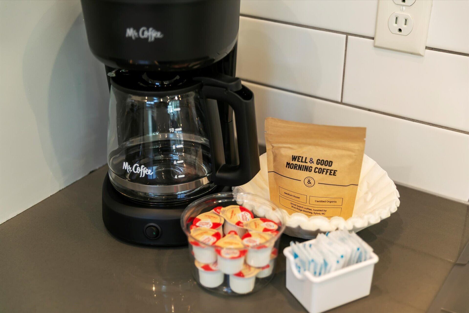 A coffee maker, a bag of Well & Good Morning Coffee, and a container of coffee pods on a kitchen counter.