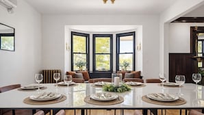 Comedor con una gran mesa dispuesta para seis personas, dos ventanas con marcos negros y una puerta de madera oscura.
