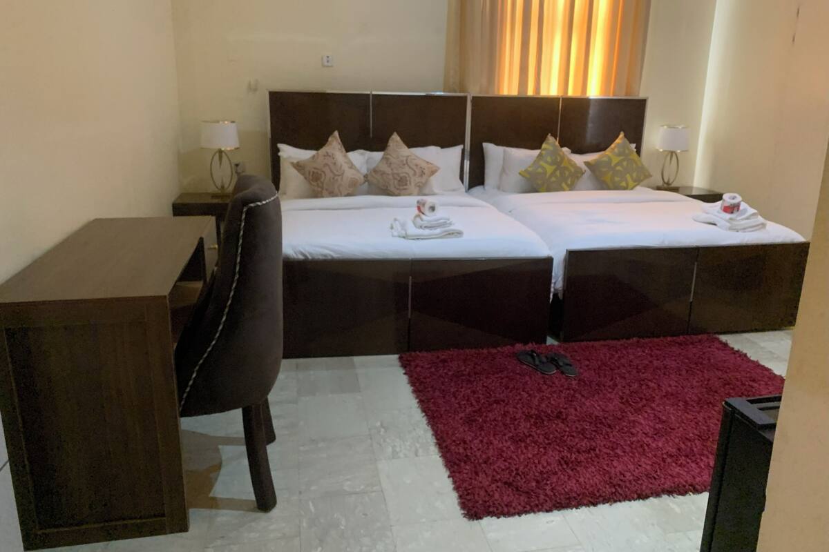 A hotel room with two beds, a desk, a chair, a red rug, and a TV.