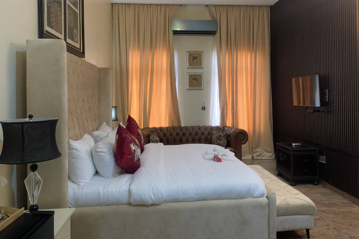 A hotel room with a large bed, a bedside table with a lamp, a brown leather sofa, a dark wooden cabinet, and a wall-mounted TV.