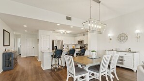 A modern kitchen with white cabinets, a central island, and a dining area with white chairs and a wooden table. - Ocean Park 118 - Hosted by Moore AI Rentals (Fernandina Beach)