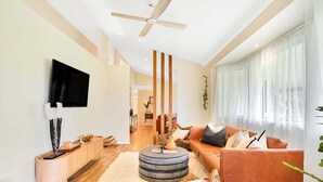 A modern living room with a flat-screen TV, a ceiling fan, a sofa, a coffee table, and a large window with sheer curtains.