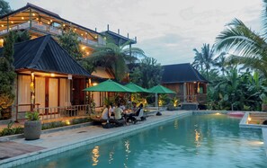 A poolside area with outdoor seating and umbrellas, surrounded by lush greenery and a building with a modern design. - Mina Baturiti Hotel & Resto (Baturiti)