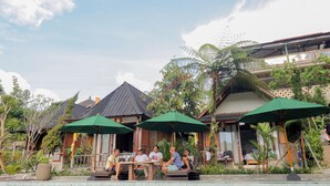 An outdoor seating area with tables and chairs under green umbrellas, surrounded by lush greenery and traditional architecture. - Mina Baturiti Hotel & Resto (Baturiti)