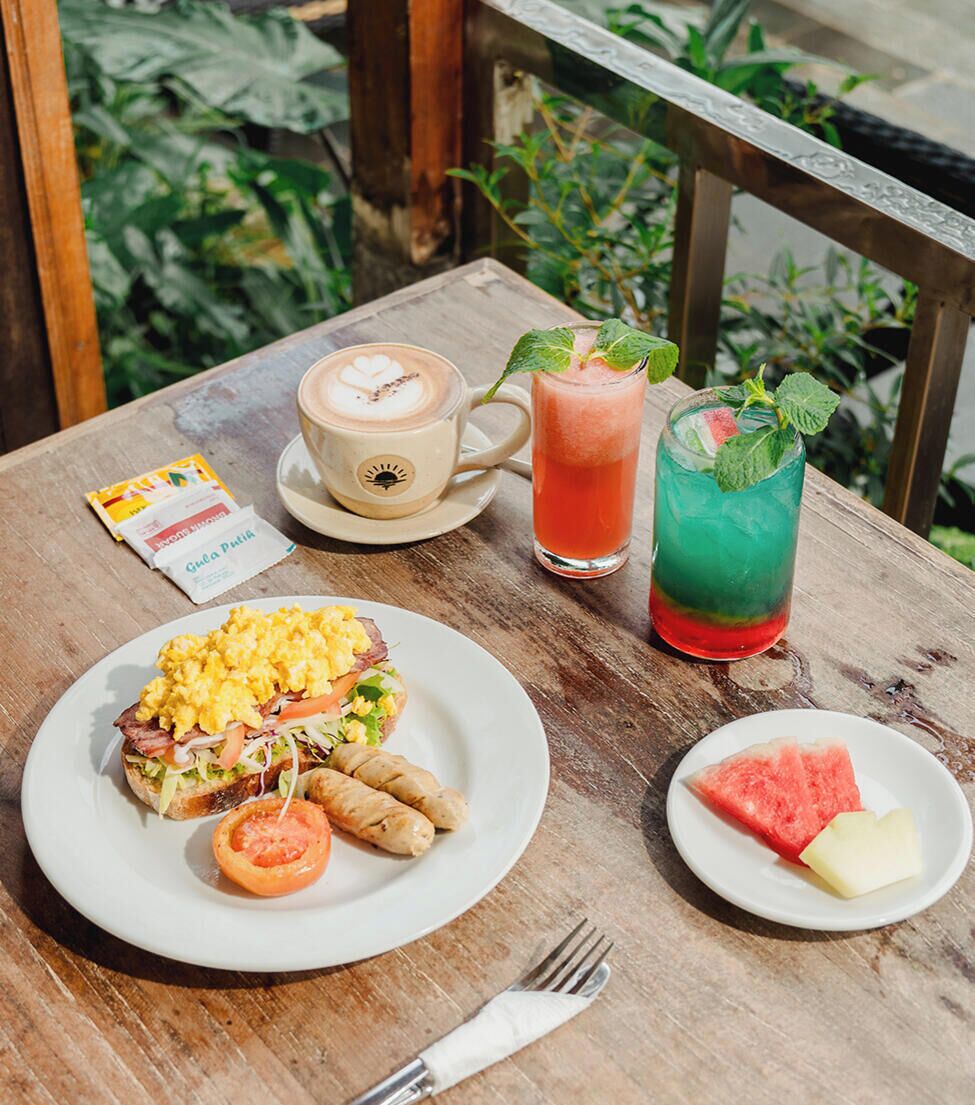 Una mesa de madera con un plato de desayuno, una taza de café, dos bebidas coloridas, un sobre de café y una rebanada de sandía.