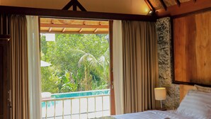 A bedroom with a bed, a nightstand, a lamp, and a view of greenery outside. - Mina Baturiti Hotel & Resto (Baturiti)