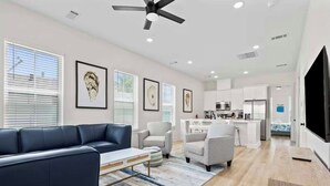 A modern living room with a sofa, armchairs, and a coffee table. There is a kitchen area with white cabinets and a dining table. - New! Galveston Retreat -Sleeps 10-EV Charger -Fast WiFi -Near Beach (Galveston)