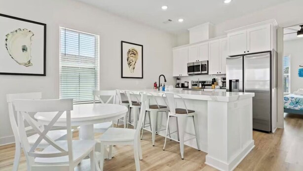 A modern kitchen with white cabinetry, a central island, and a dining area with white chairs. - New! Galveston Retreat -Sleeps 10-EV Charger -Fast WiFi -Near Beach (Galveston)