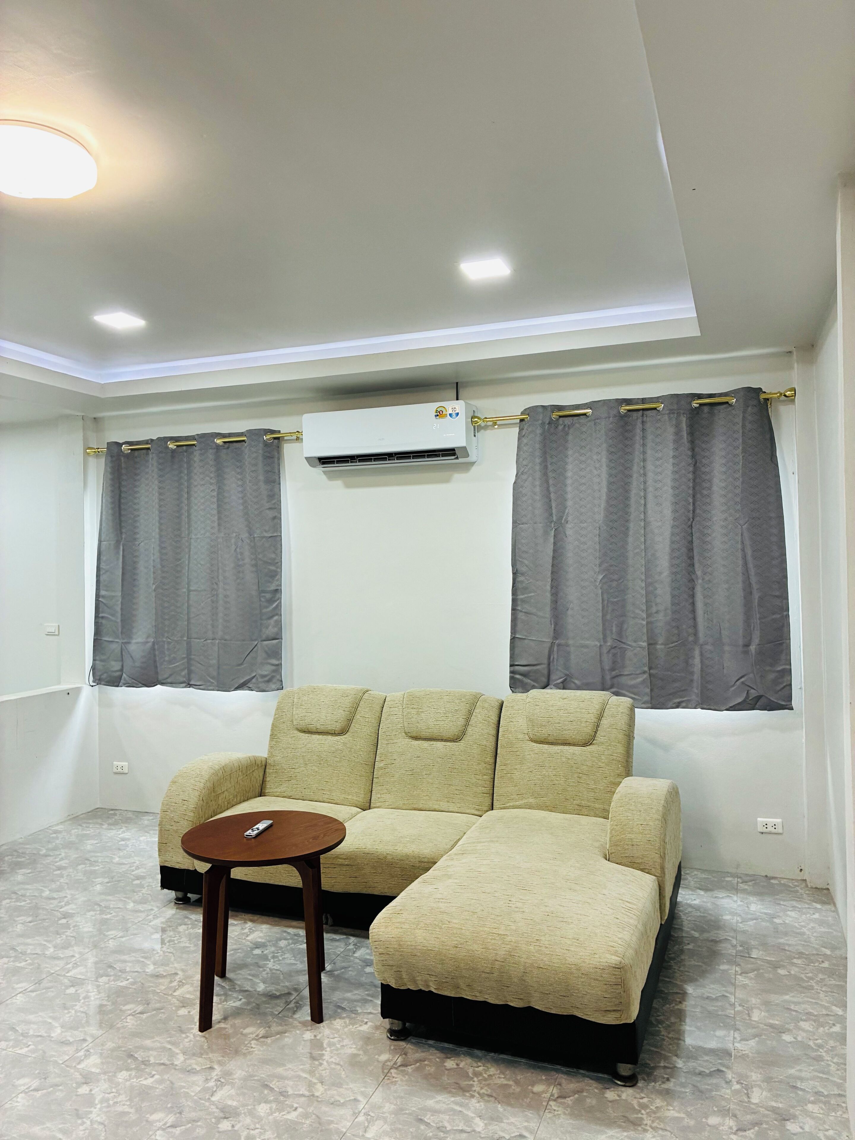 A modern living room with a beige sectional sofa, a small wooden table, and an air conditioning unit mounted on the wall.