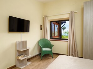 A room with a bed, a television mounted on the wall, a window with curtains, a green chair, and a small shelf with books. - Villino Sally (Piano di Sorrento)