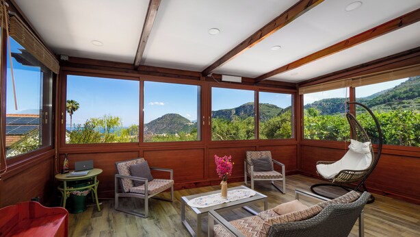 A spacious room with a wooden ceiling, large windows offering a view of green hills, and various seating options including a sofa, chairs, and a hanging chair. - Villino Sally (Piano di Sorrento)