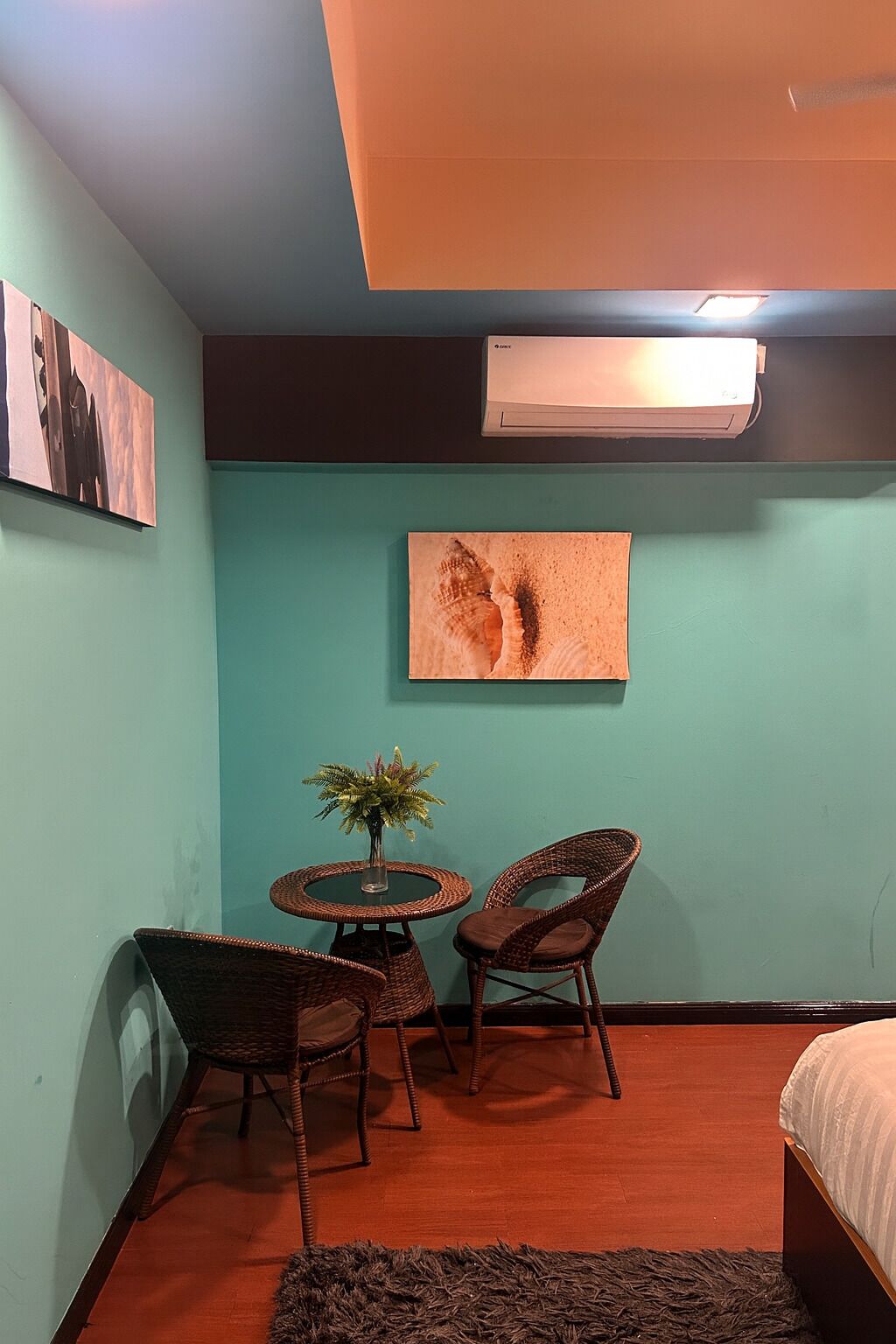 A small room with teal walls, a round table with two wicker chairs, a small plant, and a bed with a white mattress.
