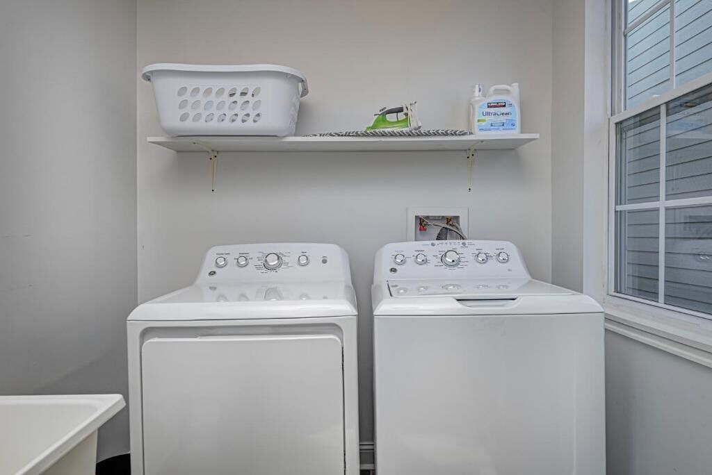 A laundry area with two white front-loading washing machines, a white laundry basket, and a shelf with a green iron and a bottle of laundry detergent.