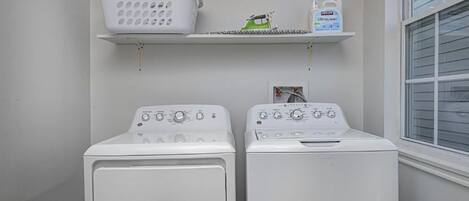 A laundry area with two white front-loading washing machines, a white laundry basket, and a shelf with a green iron and a bottle of laundry detergent.