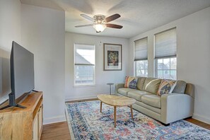 A living room with a flat-screen TV on a wooden stand, a beige sofa with patterned cushions, a wooden coffee table, a ceiling fan, and a large window with blinds. - Spacious Home for Groups with Hot Tub in Indy (Indianapolis)