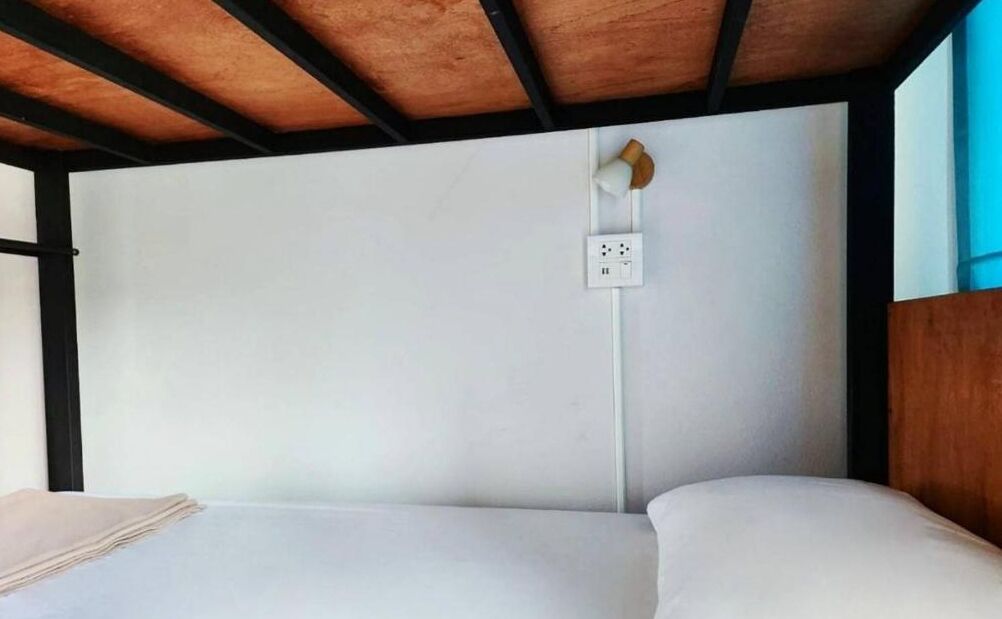 Een bed met een witte matras en een houten hoofdbord. Er is een witte muur met een stopcontact en een klein wandlampje.