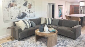 Un salon avec un canapé d’angle gris, une table basse ronde en bois et un grand tableau abstrait accroché au mur.