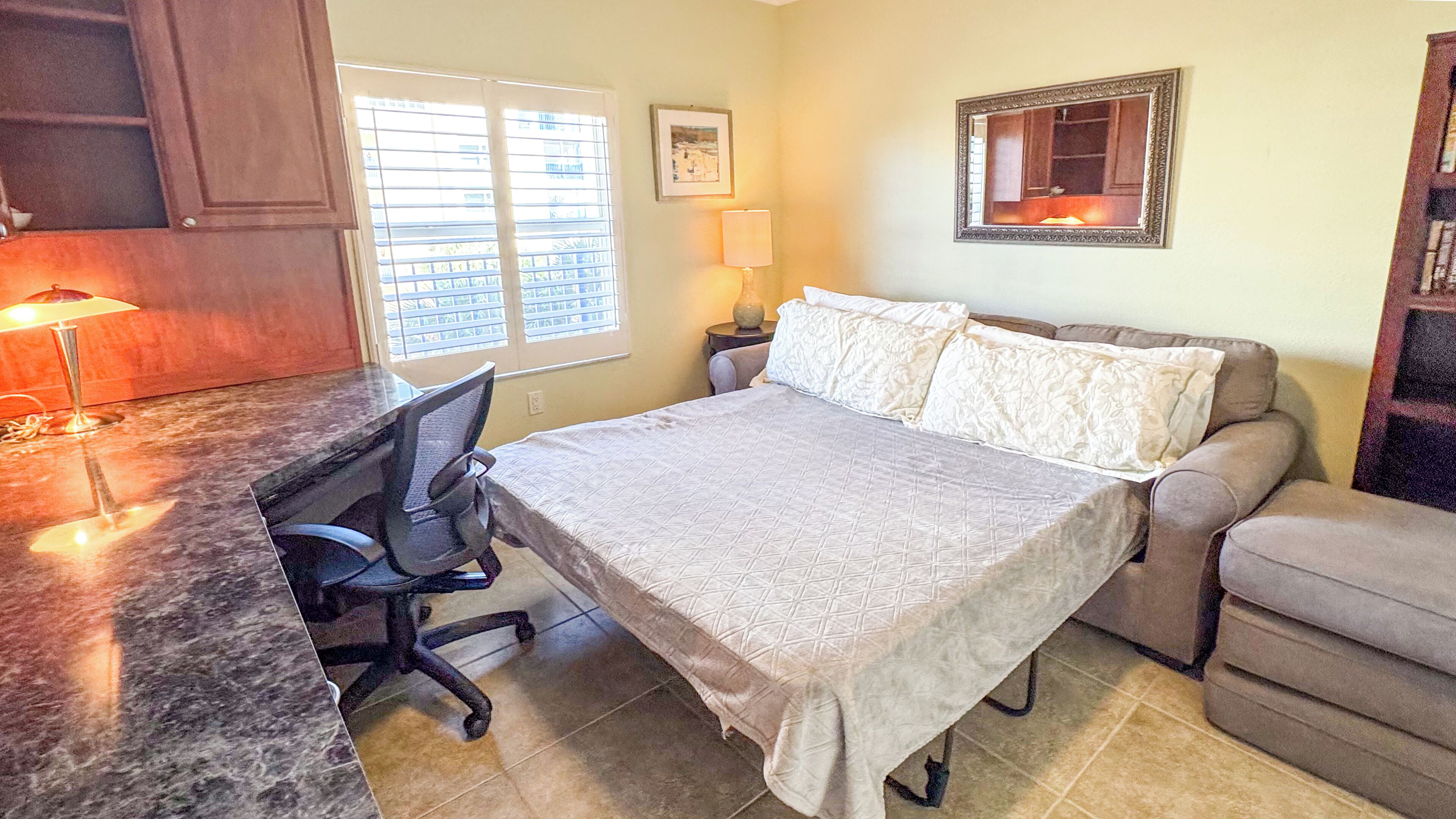 Um quarto com cama, escrivaninha, cadeira, mesa de cabeceira, luminária, janela e um quadro na parede.