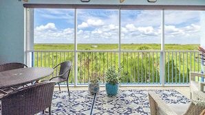 Ein Balkon mit Korbmöbeln, einem gemusterten Boden und Blick auf eine grüne Landschaft durch große Fenster.