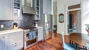 A kitchen with grey cabinets, a stainless steel stove, a refrigerator, and a dining area with a wooden table and chairs. - The Regent on Whitaker | Downtown Savannah Luxury (Savannah)