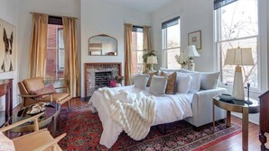 A bedroom with a bed, a chair, a table, a fireplace, and large windows. - The Regent on Whitaker | Downtown Savannah Luxury (Savannah)