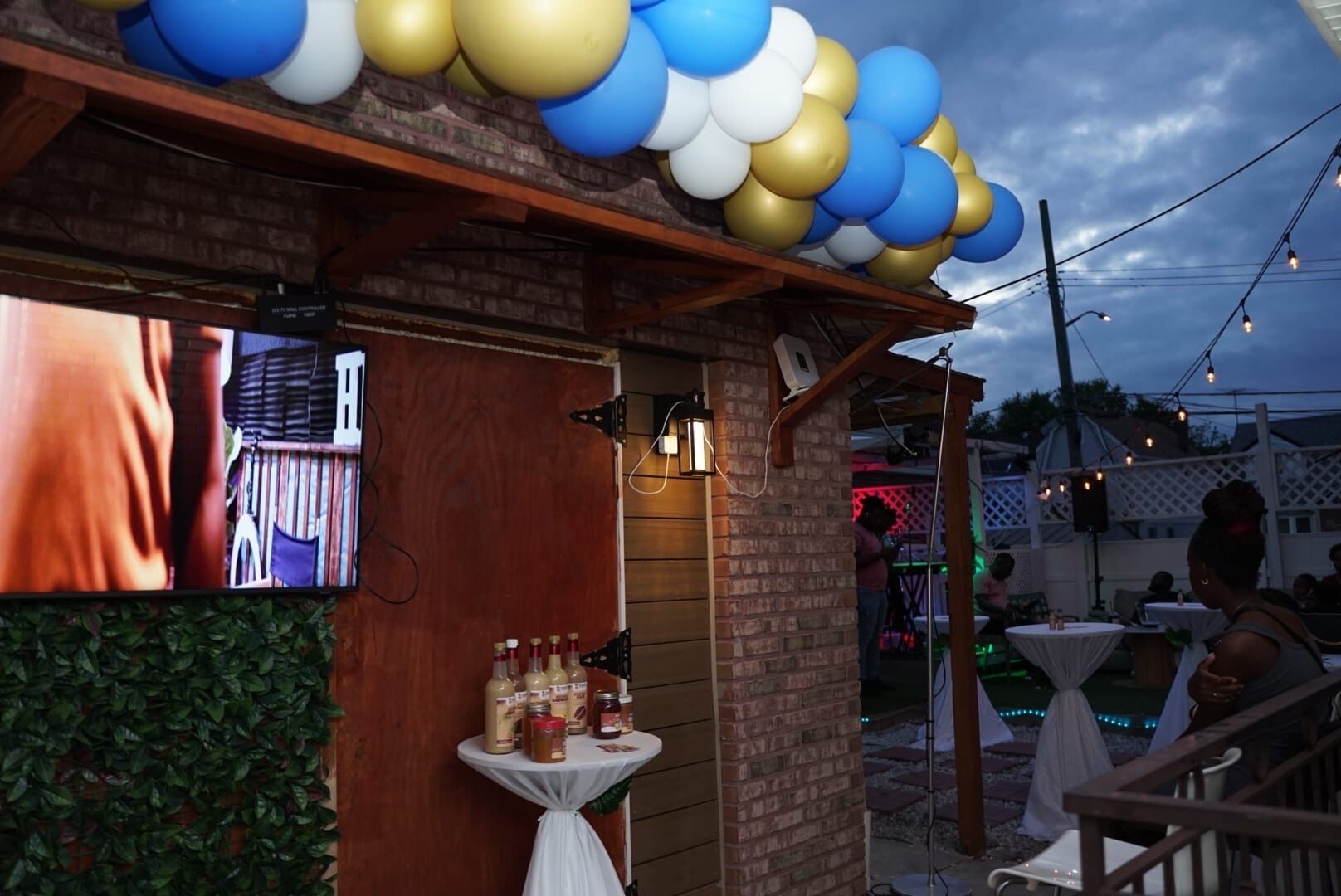 Un espace extérieur aménagé en patio, avec des murs en briques, une pergola en bois et des guirlandes lumineuses. On y trouve des tables et des chaises disposées pour les repas, ainsi qu’une petite table sur laquelle sont placés des condiments.