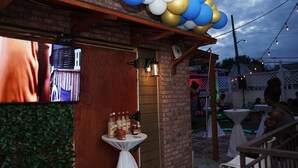 Un patio al aire libre con paredes de ladrillo, una pérgola de madera y luces colgantes. Hay mesas y sillas dispuestas para comer, y una pequeña mesa con salsas y aderezos.