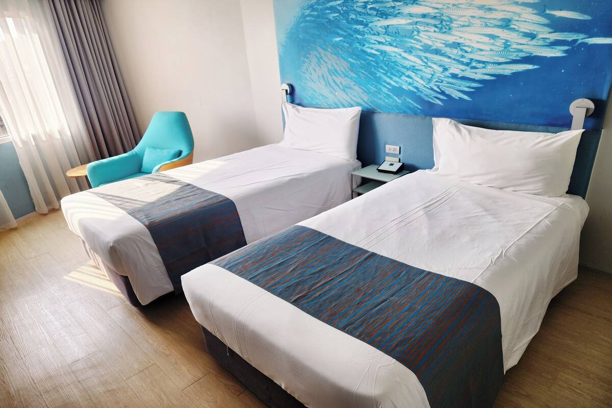 Een hotelkamer met twee bedden, een nachtkastje, een stoel en een groot schilderij aan de muur.