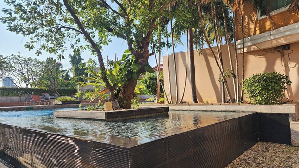 A swimming pool with a black textured edge, surrounded by greenery and a beige building. - Bhanwar Singh Palace Pattaya (Pattaya)