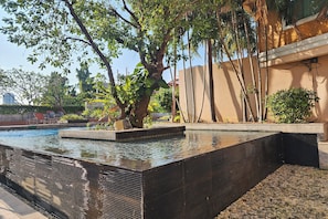 En swimmingpool med sort, struktureret kant, omgivet af grøn vegetation og en beige bygning.