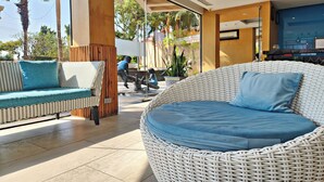 A wicker lounge area with a blue cushion, a wicker sofa with a blue cushion, and a view of the outdoors including palm trees and a person walking. - Bhanwar Singh Palace Pattaya (Pattaya)
