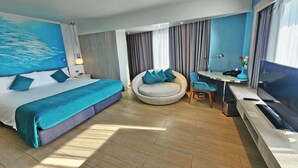 A modern hotel room with a large bed, a round seating area, a desk with a TV, and a large window with curtains. - Bhanwar Singh Palace Pattaya (Pattaya)