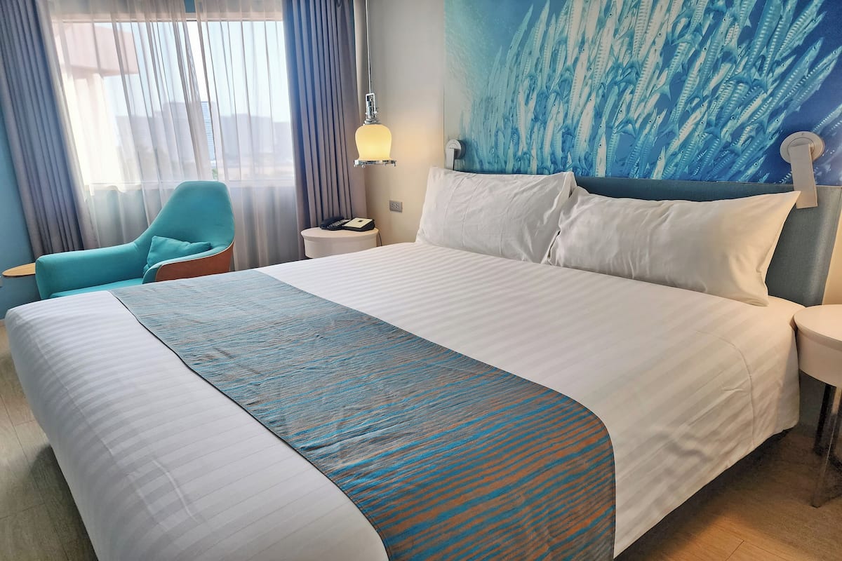Hotelkamer met een groot bed, een nachtkastje, een lamp, een stoel en een muur met een blauw abstract ontwerp.