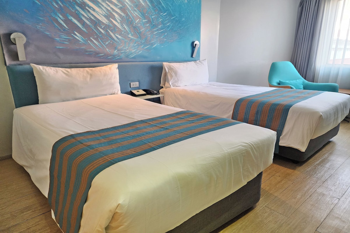 Een hotelkamer met twee bedden, een blauwe stoel en een groot abstract schilderij aan de muur.