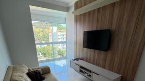 Una sala de estar moderna con un sofá seccional color beige, un televisor montado y un amplio ventanal con vistas a edificios y áreas verdes.