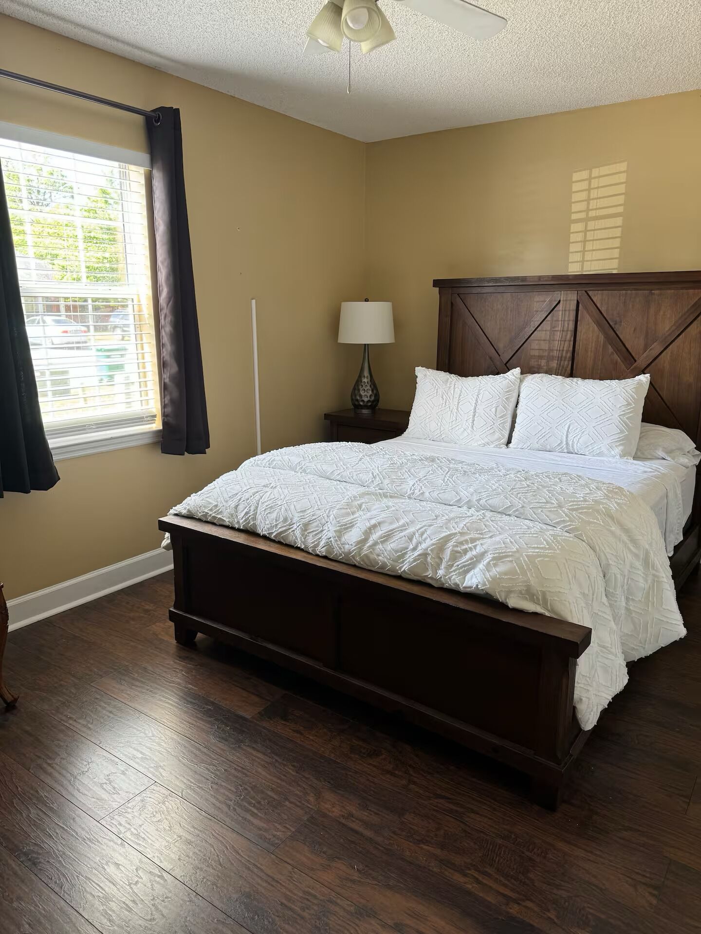 Un dormitorio con cama de madera, mesita de noche con lámpara, ventilador de techo y ventana con cortinas.