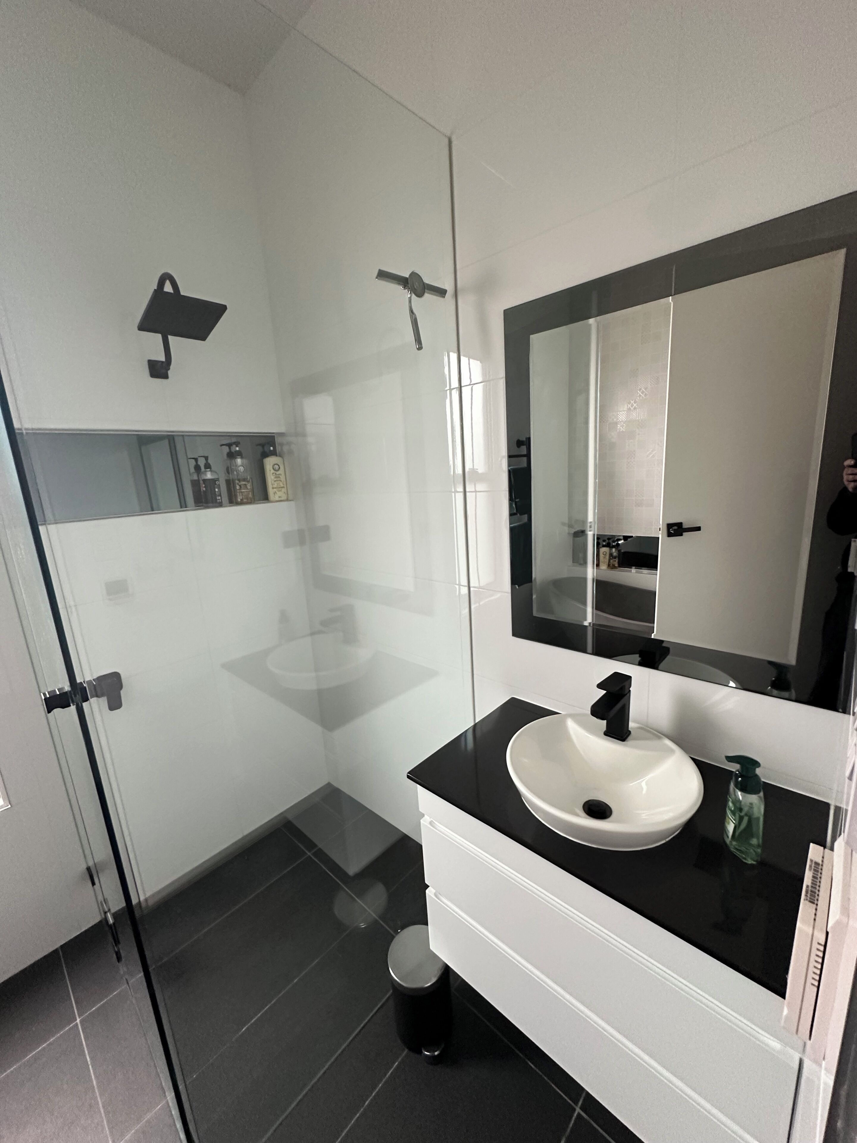 Ein modernes Badezimmer mit einer Duschkabine aus Glas, einem weißen Waschbecken mit schwarzer Arbeitsplatte und einem großen Spiegel darüber.